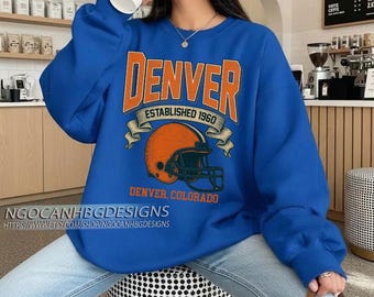May include: A vibrant blue sweatshirt with the word "DENVER" in orange lettering, and "ESTABLISHED 1960" above an orange football helmet graphic. Below the helmet, the text reads "DENVER, COLORADO". The shop's name and website are at the bottom.
