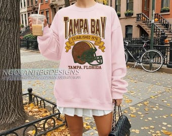 Puede incluir: Una sudadera rosa claro con el texto "TAMPA BAY" en una fuente desgastada, un gráfico de casco de fútbol americano y "TAMPA, FLORIDA" debajo. La sudadera también tiene el texto "ESTABLISHED 1976".