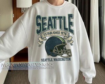 May include: White sweatshirt with a graphic design featuring the word "SEATTLE" in green and blue, the text "ESTABLISHED 1976", and a football helmet. The text "SEATTLE, WASHINGTON" is also included.