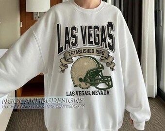 May include: White sweatshirt with a graphic design featuring "LAS VEGAS" in bold black letters above a football helmet. The design includes the text "ESTABLISHED 1960" and "LAS VEGAS, NEVADA". The sweatshirt has a crew neck and long sleeves.