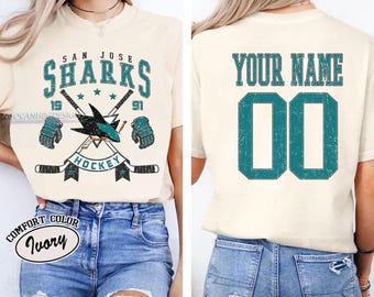 May include: A white t-shirt with a vintage-style San Jose Sharks hockey logo on the front and the text "Your Name 00" on the back. The shirt is available in ivory.