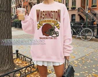 Op de afbeelding: Een lichtroze sweatshirt met een afbeelding van een Washington D.C. footballhelm. De afbeelding bevat het woord "WASHINGTON" in bordeauxrode en gouden letters, met "ESTABLISHED 1932" en "WASHINGTON, D.C." eronder.