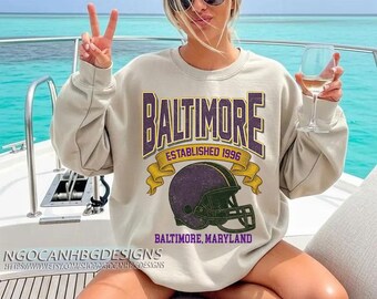 Puede incluir: Una sudadera beige con la palabra "BALTIMORE" en letras moradas, un casco de fútbol americano morado y el texto "ESTABLISHED 1996" y "BALTIMORE, MARYLAND". La sudadera la lleva una persona en un barco.