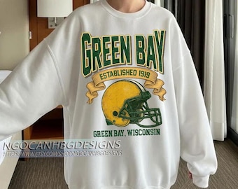 Peut inclure: Sweat-shirt blanc avec un motif des Packers de Green Bay. Le motif comprend les mots "GREEN BAY" en vert et or, un casque de football américain, et le texte "ESTABLISHED 1919" et "GREEN BAY, WISCONSIN".