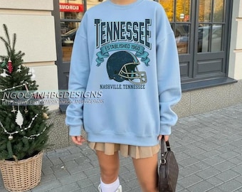 Op de afbeelding: Een lichtblauwe sweatshirt met het woord "TENNESSEE" in een donkergroene lettertype, samen met een afbeelding van een footballhelm. De tekst "ESTABLISHED 1960" en "NASHVILLE, TENNESSEE" zijn ook afgebeeld. Kledingstuk.