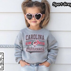 May include: A young child wearing a grey sweatshirt with the Carolina Hurricanes logo and the text "CAROLINA EST. 1997 Hurricanes". The child is wearing pink heart-shaped sunglasses.