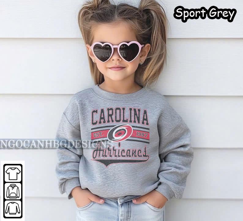 May include: A young child wearing a grey sweatshirt with the Carolina Hurricanes logo and the text "CAROLINA EST. 1997 Hurricanes". The child is wearing pink heart-shaped sunglasses.