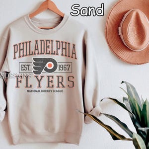 Op de afbeelding: Een zandkleurig sweatshirt met de tekst "Philadelphia Flyers" en het teamlogo. Het logo is een gestileerde zwarte en oranje letter "P" met een hockeystick en een puck. De tekst "Est. 1967" staat onder het logo. De tekst "National Hockey League" staat onder de tekst "Flyers".