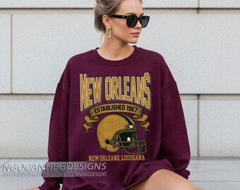 May include: A maroon sweatshirt with the words "NEW ORLEANS" in gold lettering, along with a gold football helmet graphic. The text "ESTABLISHED 1967" and "NEW ORLEANS, LOUISIANA" are also displayed. The sweatshirt is a crew neck style.
