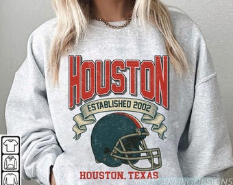 Può includere: Felpa grigio chiaro con grafica "Houston" rossa e bianca invecchiata, "Established 2002" e un casco da football americano. Il testo "Houston, Texas" è sotto.