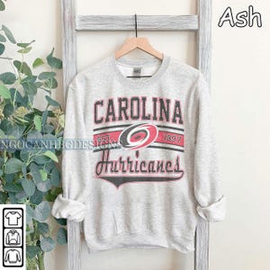 May include: A gray sweatshirt with a vintage Carolina Hurricanes logo. The logo features the team's name, a red and black hurricane symbol, and the text "EST. 1997".