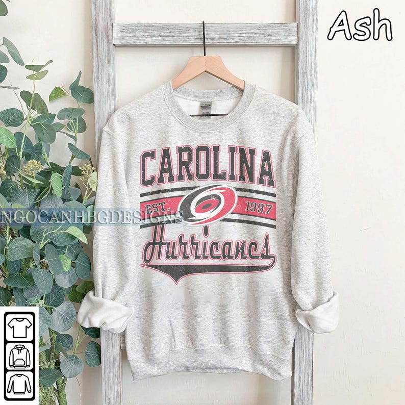 May include: A gray sweatshirt with a vintage Carolina Hurricanes logo. The logo features the team's name, a red and black hurricane symbol, and the text "EST. 1997".