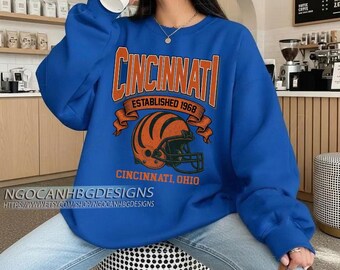 以下が含まれることがあります： オレンジ色の文字で「CINCINNATI」と書かれた、ロイヤルブルーのスウェットシャツ。「ESTABLISHED 1968」はオレンジ色のフットボールヘルメットのグラフィックの上にあります。ヘルメットの下には「CINCINNATI, OHIO」と書かれています。