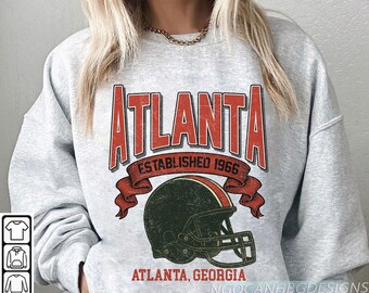 Puede incluir: Sudadera gris claro con la palabra "ATLANTA" en rojo, "ESTABLISHED 1966" y un diseño de casco de fútbol americano. El texto "ATLANTA, GEORGIA" está debajo. El diseño tiene un aspecto vintage.