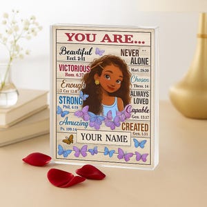 Puede incluir: Un bloque acrílico decorativo con una ilustración de dibujos animados de una niña con cabello castaño y un vestido azul. El bloque tiene texto inspirador y gráficos de mariposas. Las palabras "YOU ARE..." están en la parte superior, con frases como "Beautiful" y "Victorious".