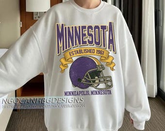 Puede incluir: Sudadera blanca con un diseño morado y dorado. El diseño incluye la palabra "Minnesota", un casco de fútbol americano y el texto "Established 1961" y "Minneapolis, Minnesota". La sudadera tiene cuello redondo.