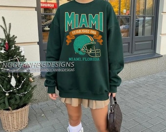 Könnte beinhalten: Ein dunkelgrünes Sweatshirt mit dem Wort "MIAMI" in türkisfarbenen Buchstaben, einer türkisfarbenen Footballhelm-Grafik und dem Text "ESTABLISHED 1966" und "MIAMI, FLORIDA". Das Sweatshirt wird mit einem Faltenrock und weißen Stiefeln getragen.