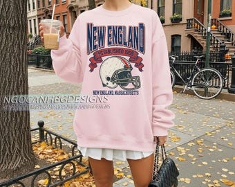 Könnte beinhalten: Ein hellrosa Sweatshirt mit einem Marineblau "New England" Grafik, einem Footballhelm und dem Text "Established 1959". Das Sweatshirt ist Teil einer Bekleidungskollektion.