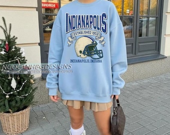 May include: A light blue sweatshirt with the word "Indianapolis" in navy blue arched across the top. Below the word is "Established 1953" and a football helmet graphic. The text "Indianapolis, Indiana" is below the helmet.