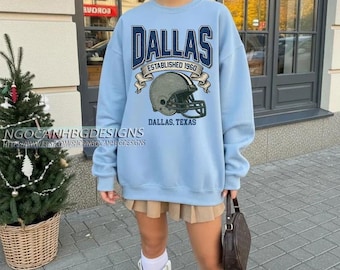 May include: A pale blue sweatshirt featuring the word "DALLAS" in navy blue lettering, a football helmet graphic, and the text "ESTABLISHED 1960" and "DALLAS, TEXAS". The sweatshirt is worn with a pleated skirt and white boots.