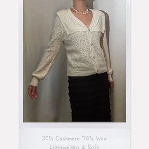 Puede incluir: Un cárdigan de color crema de manga larga con cuello ancho, cierre de botones frontal y un intrincado patrón de punto calado. La prenda está etiquetada como 30% Cashmere 70% Wool, Lightweight & Soft. Se combina con una falda negra y un collar de cuentas.