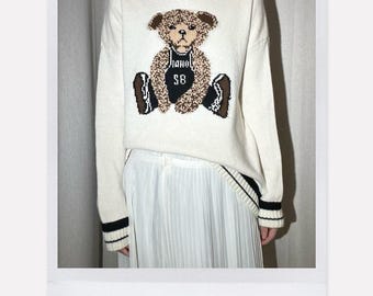 May include: Cream-colored sweater with a teddy bear graphic. The bear has a brown and tan pattern, wearing a black jersey with "SB" on it. The sweater has black and white striped trim and the text "100% Wool".
