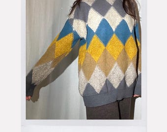 May include: A long-sleeve turtleneck sweater featuring a multi-colored argyle diamond pattern in shades of grey, blue, yellow, and beige. The knit fabric has a textured appearance, and the text 100% Cashmere is printed below the garment.
