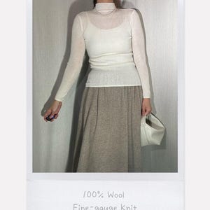May include: A sheer white long-sleeve mock neck top layered over a white camisole, paired with a beige midi skirt and a white handbag. The text 100% Wool Fine-gauge Knit is visible at the bottom. The outfit includes a simple pearl necklace.
