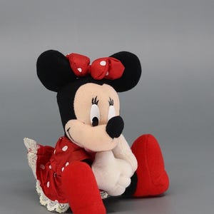 May include: A plush Minnie Mouse doll, featuring black ears, a red polka-dot bow, and a red dress with white polka dots. The doll has white gloves and red shoes. The doll is sitting down.