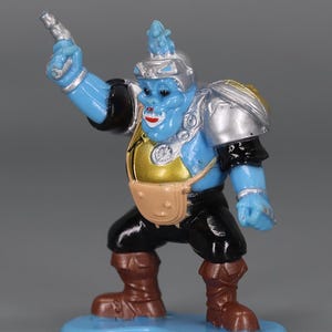 May include: A blue action figure with silver armor, a gold chest, and a brown satchel. The figure is holding a silver gun and is wearing black pants and brown boots. It stands on a blue base.