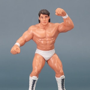 May include: A plastic figurine of a muscular wrestler in a dynamic pose. The figure is wearing white briefs, wristbands, and boots. The wrestler has dark hair and is flexing his arm. The background is a solid grey.