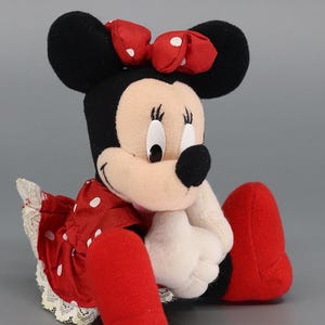 May include: A plush Minnie Mouse doll, featuring black ears, a red bow with white polka dots, and a red dress with white polka dots. The doll has white gloves and red shoes. The doll is sitting down.