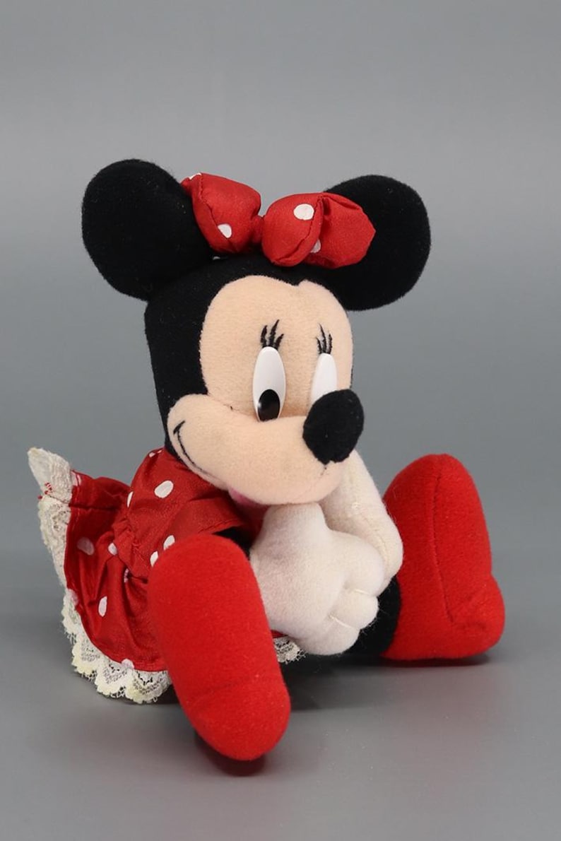May include: A plush Minnie Mouse doll, featuring black ears, a red bow with white polka dots, and a red dress with white polka dots. The doll has white gloves and red shoes. The doll is sitting down.