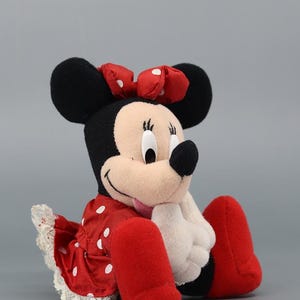 May include: A plush Minnie Mouse doll, featuring black ears, a red polka-dot dress, and a matching bow. The doll has white gloves and red shoes. The overall design is a classic representation of the iconic cartoon character.