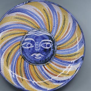 May include: A decorative ceramic plate with a sun face design. The plate has a blue face with white eyes and a mouth. The background has a spiral pattern in yellow, blue, and brown. The plate is round.
