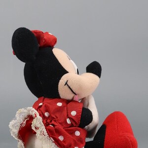 May include: A plush Minnie Mouse doll, featuring black ears, a red bow, and a red dress with white polka dots. The dress has a white lace trim. The doll's feet are red, and it is lying on its side.