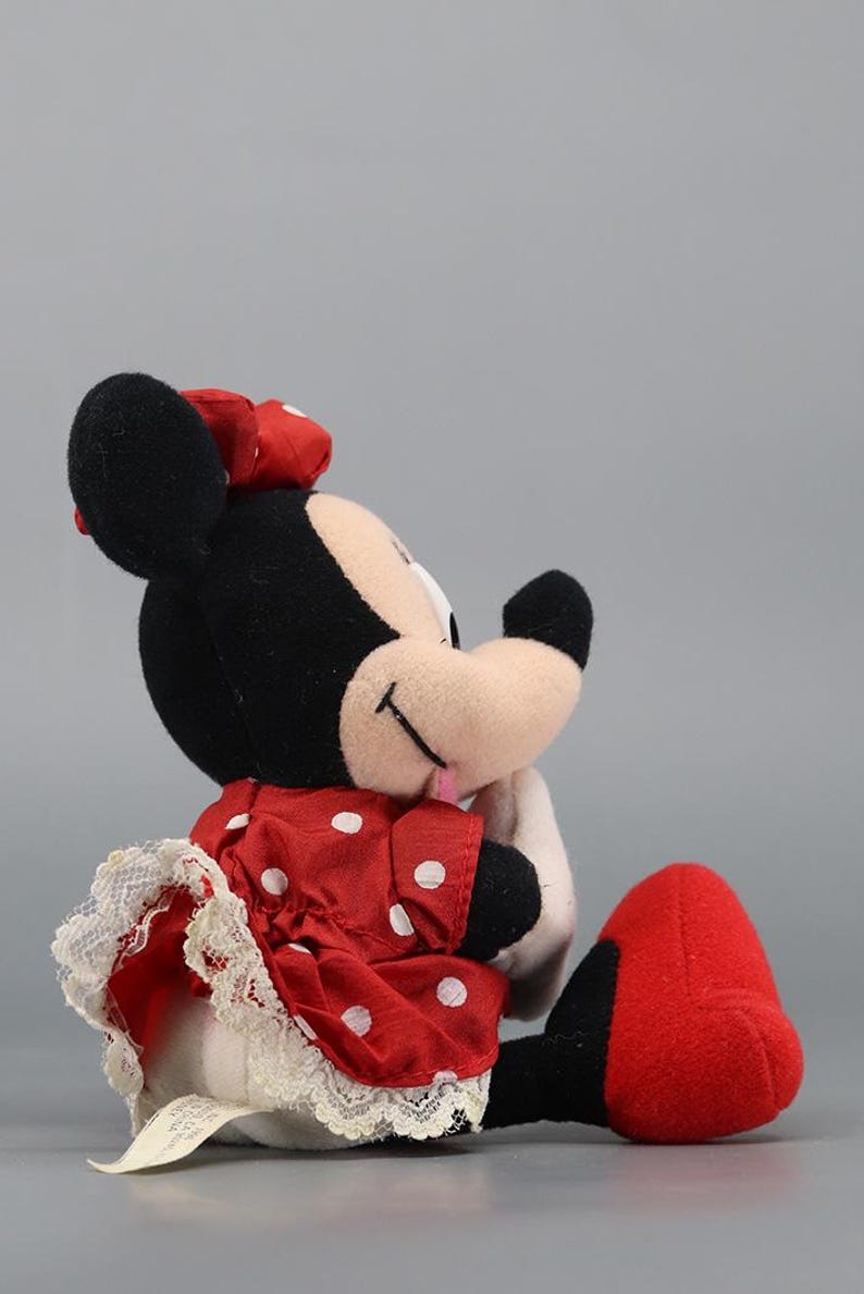 May include: A plush Minnie Mouse doll, featuring black ears, a red bow, and a red dress with white polka dots. The dress has a white lace trim. The doll's feet are red, and it is lying on its side.