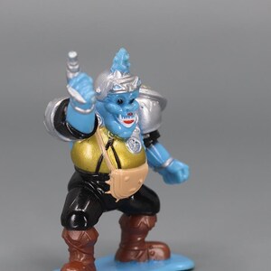 May include: A blue action figure of a fantasy creature with a silver helmet and shoulder pads. The figure has a gold torso, black pants, and brown boots. It stands on a light blue base, holding a silver weapon.