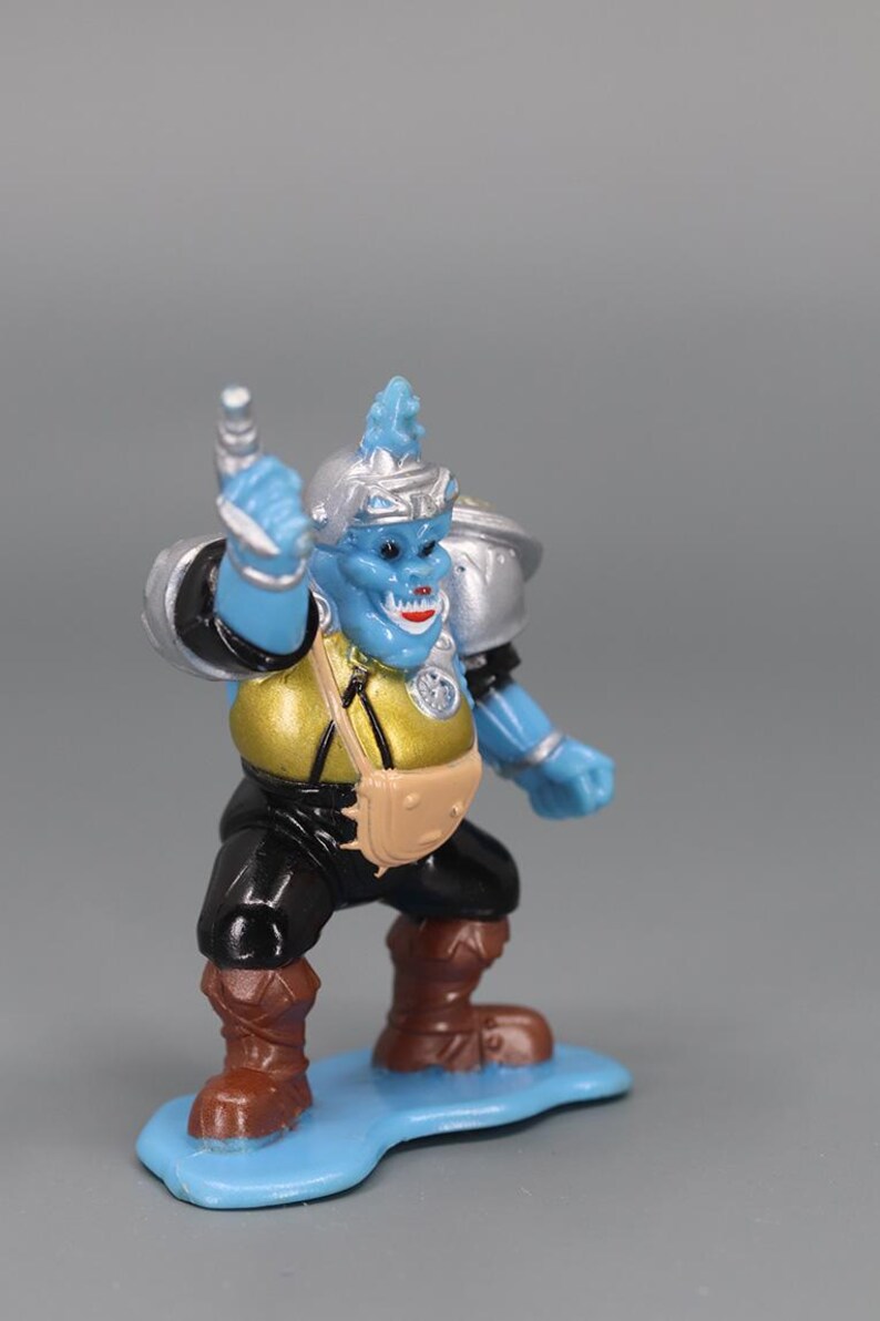 May include: A blue action figure of a fantasy creature with a silver helmet and shoulder pads. The figure has a gold torso, black pants, and brown boots. It stands on a light blue base, holding a silver weapon.