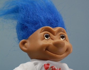 May include: A blue haired troll doll wearing a white shirt with a red heart and the text "Papa Knoll" on it. The doll is wearing blue denim pants.