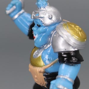 May include: A blue action figure with silver armor, a silver helmet, and a gold chest plate. The figure has a blue face with red lips and a brown belt. The figure is in a dynamic pose, with one arm raised.