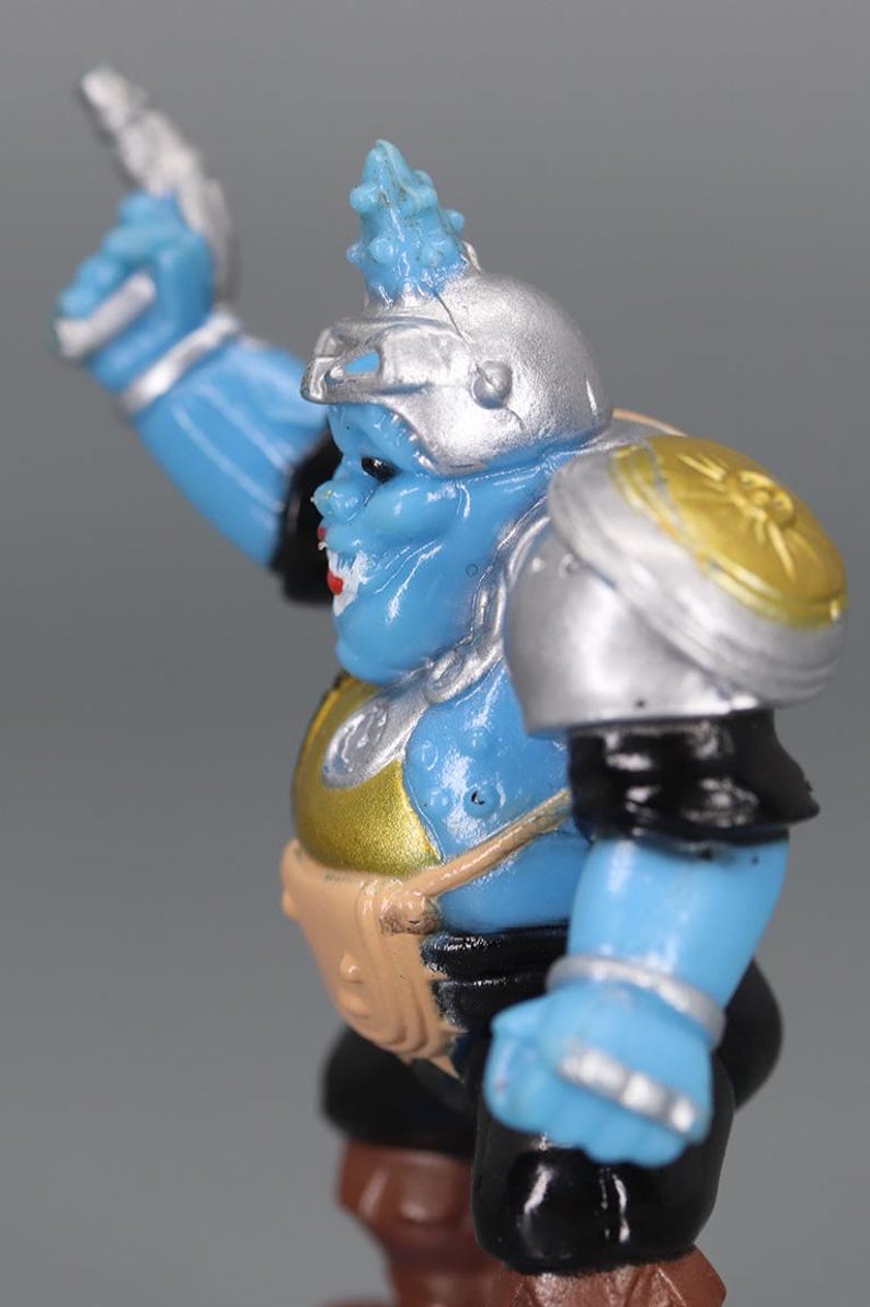 May include: A blue action figure with silver armor, a silver helmet, and a gold chest plate. The figure has a blue face with red lips and a brown belt. The figure is in a dynamic pose, with one arm raised.