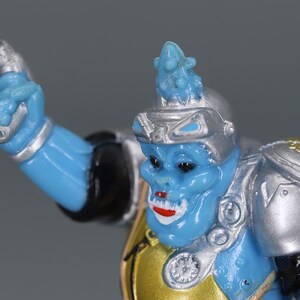 May include: A blue action figure with a silver helmet and shoulder pads. The figure has a gold chest plate and a tan belt. The figure's face has red lips and a small blue headpiece.