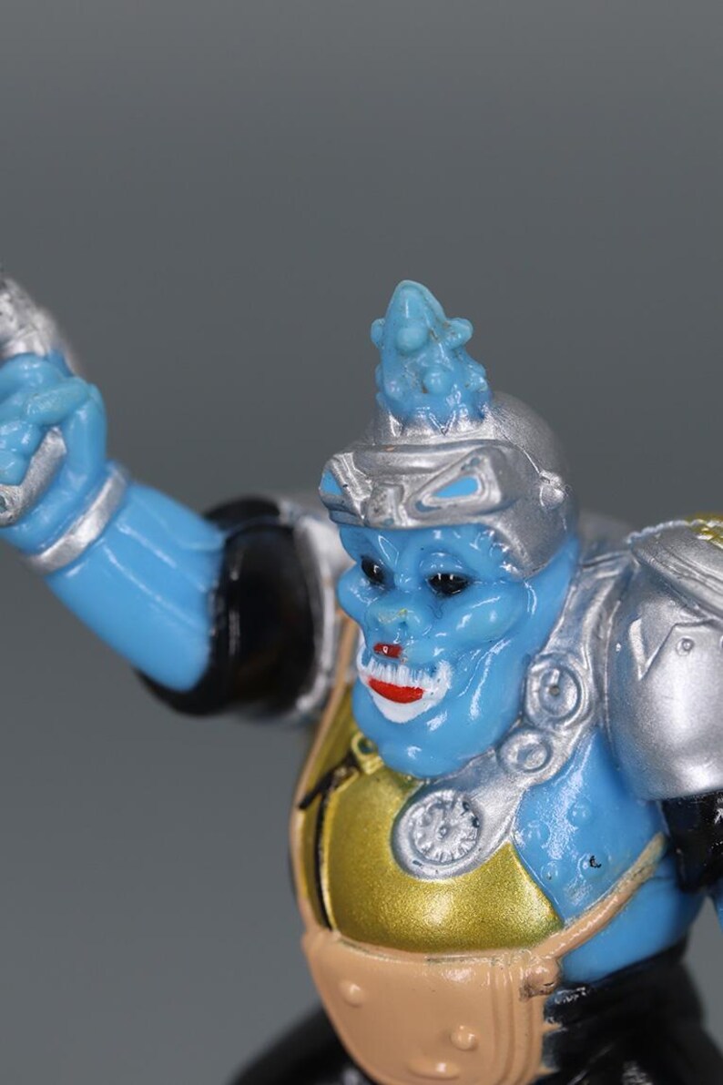 May include: A blue action figure with a silver helmet and shoulder pads. The figure has a gold chest plate and a tan belt. The figure's face has red lips and a small blue headpiece.