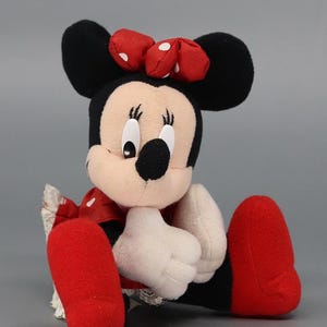 May include: A plush Minnie Mouse doll, featuring black ears, a red bow with white polka dots, and a smiling face. The doll wears a red dress with white polka dots, white gloves, and red shoes. The doll is sitting.