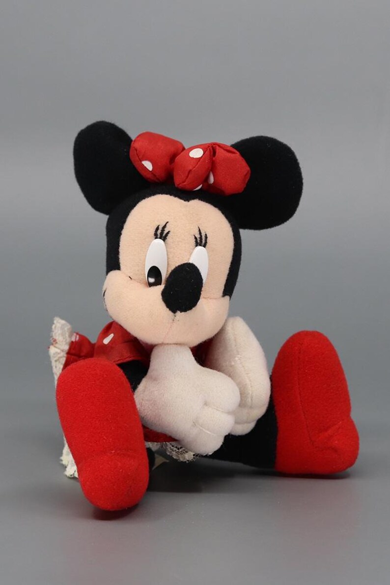 May include: A plush Minnie Mouse doll, featuring black ears, a red bow with white polka dots, and a smiling face. The doll wears a red dress with white polka dots, white gloves, and red shoes. The doll is sitting.
