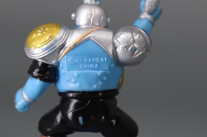 May include: A blue and silver action figure with gold accents. The figure has a helmet, shoulder pads, and black pants. The text "&copy;93 BANDAI CHINA" is visible on the back.