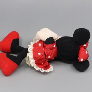 May include: A plush Minnie Mouse doll, lying on its side, features a black head with round ears and a red bow with white polka dots. The doll wears a red dress with white polka dots and red shoes.