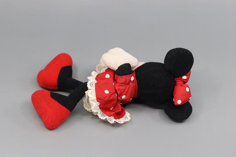 May include: A plush Minnie Mouse doll, lying on its side, features a black head with round ears and a red bow with white polka dots. The doll wears a red dress with white polka dots and red shoes.