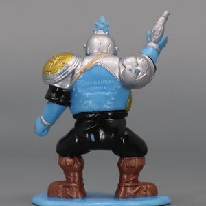 May include: A blue, silver, and black action figure with a gun raised in the air. The figure has brown boots, black pants, and a blue torso. The figure is standing on a light blue base. The figure has gold and silver shoulder pads.